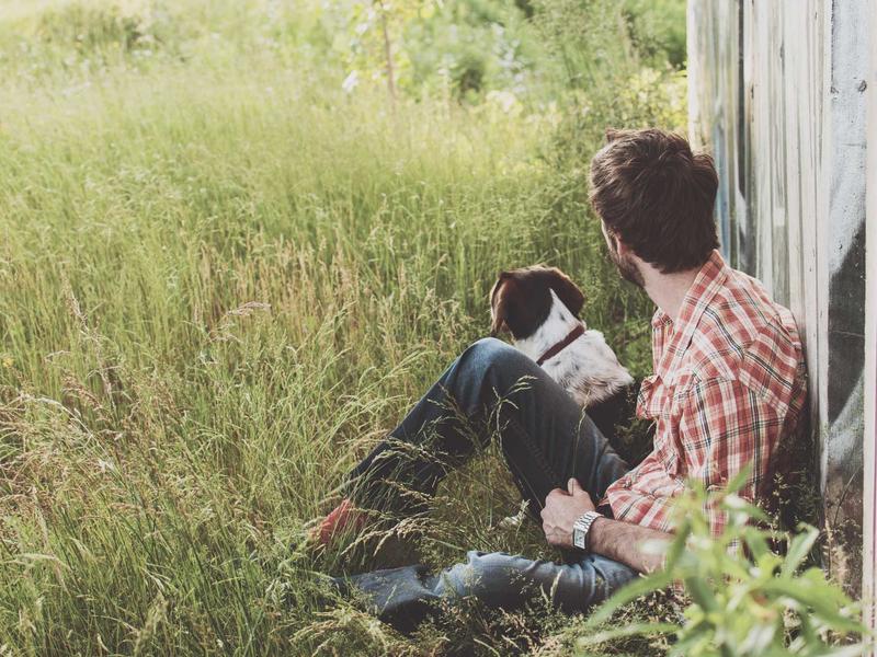 Person sitting in tall grass next to a dog beside a weathered building