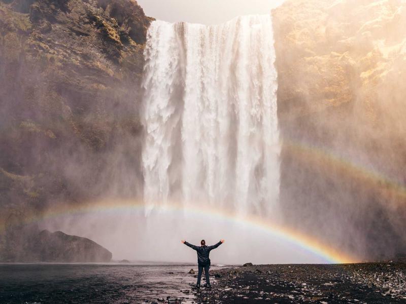 Person standing with arms outstretched before a towering waterfall with a rainbow in the mist