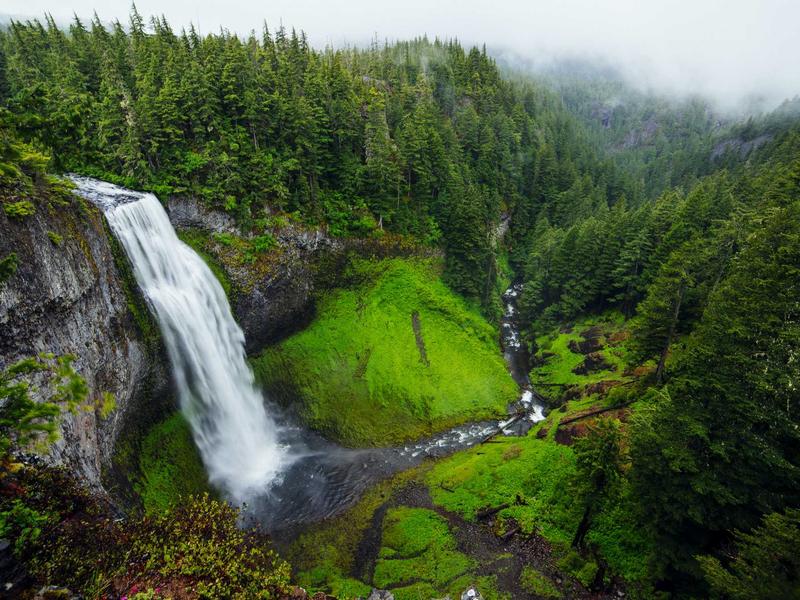 Waterfall cascading off a cliff into a lush green valley shrouded in fog